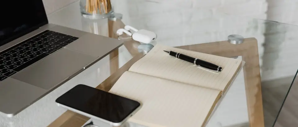 A laptop, a phone and an open notebook on a glass desk