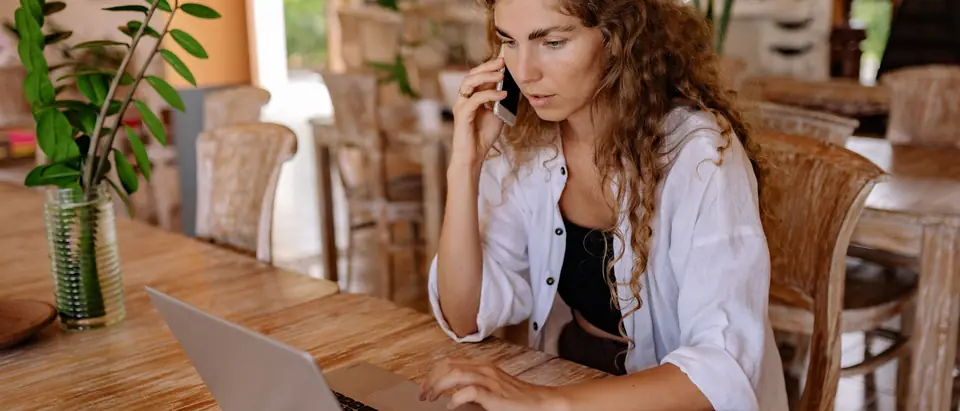 A woman on a phone call at a laptop on a wooden table