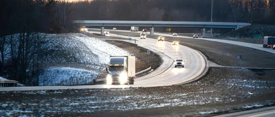 Headlights on a curving interstate at dusk beside snowy banks